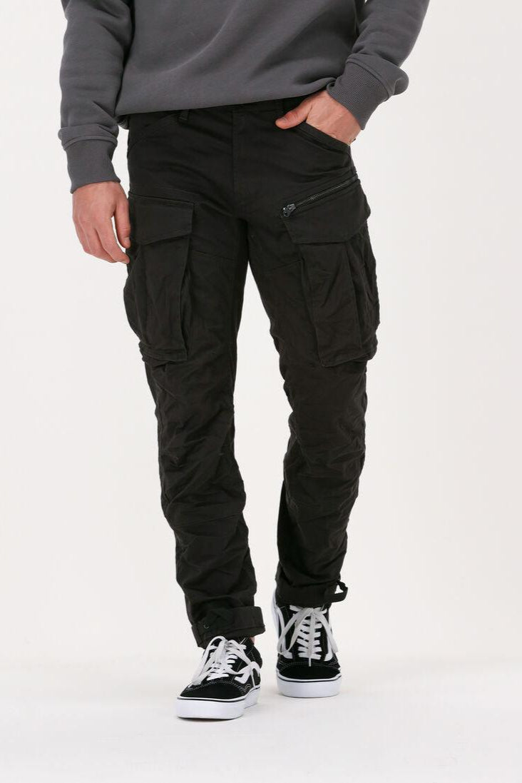 A person standing against a white background, wearing green regular fit cargo pants with multiple pockets and metallic zip detailing.