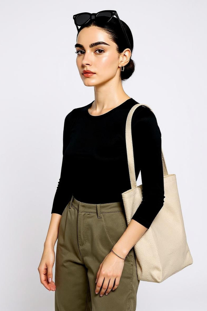Woman wearing a black top and green pants, holding a beige tote bag against a white background