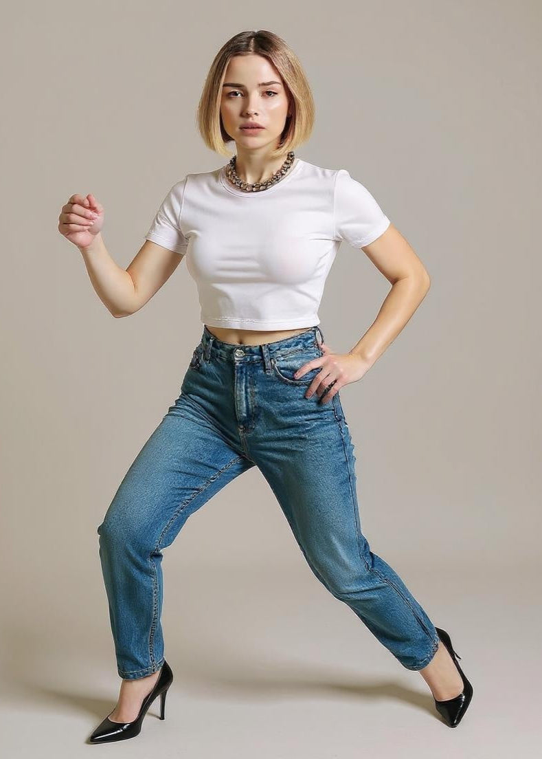 Woman wearing a white crop top and blue jeans posing against a plain background