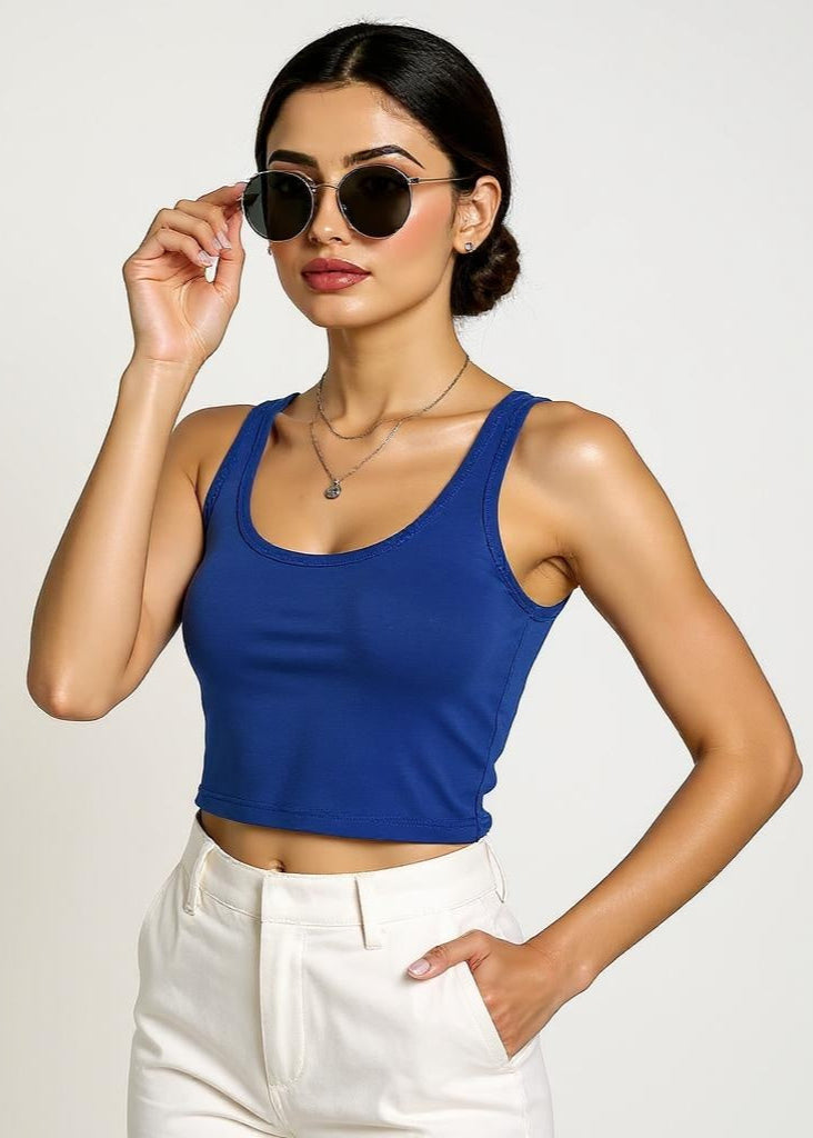 Woman wearing a blue tank top and white pants, holding sunglasses against a plain background