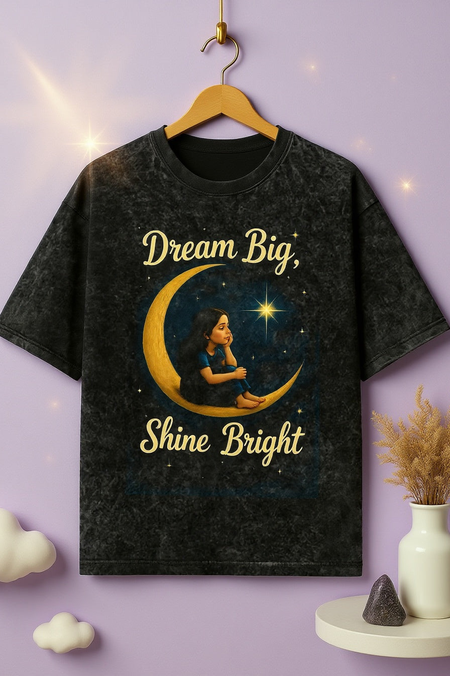 Black t-shirt with 'Dream Big, Shine Bright' design on a hanger against a purple background.