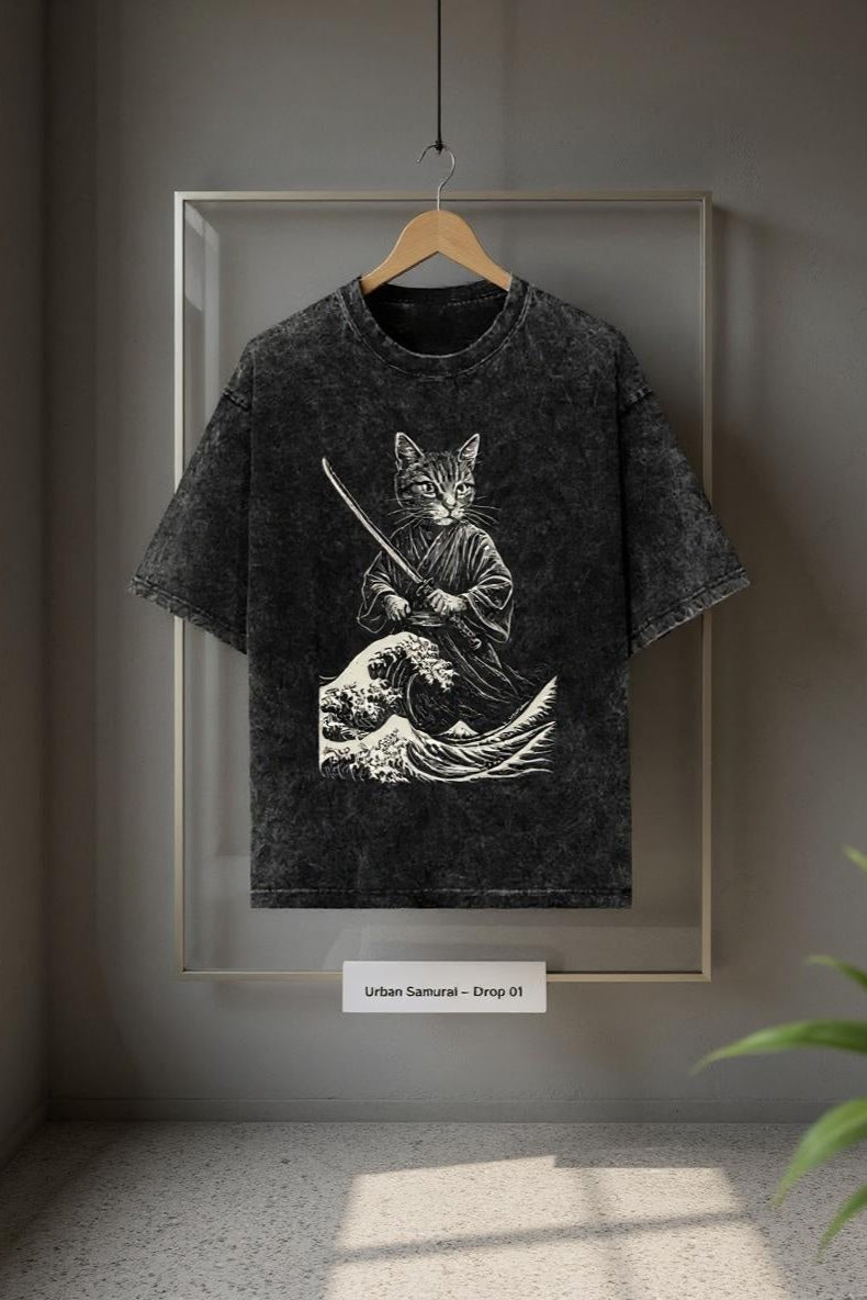 T-shirt with a graphic design of a cat in a samurai outfit on a hanger against a gray wall.